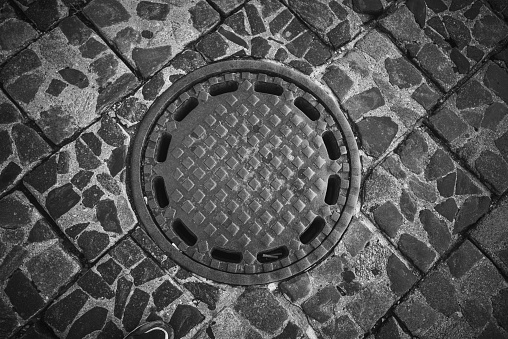 A metal round sewer manhole on the pavement of a pedestrian city street. Black and white image.