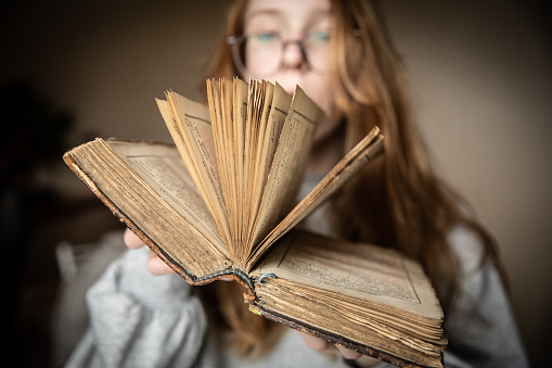 An open vintage book in the hands of a cute teenage girl in glasses and with red hair, in a semi-dark room illuminated by the light from the window. High quality photo.