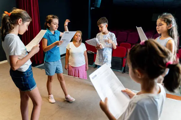 Photo of Group of children enjoying drama club rehearsal. They are reading script with their drama teacher.
