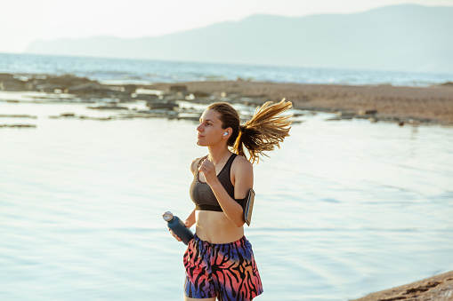 A young Caucasian woman is running on  the beach on a sunny morning and listening to music.