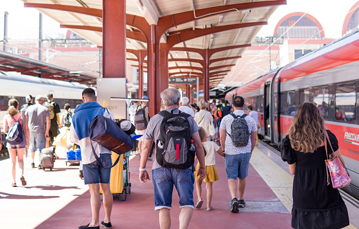 In the middle of a Train Station, a Group of Tourists Walks through the Platform with their Suitcases and Backpacks ready to Enjoy their Holidays and Meet their Friends and Family