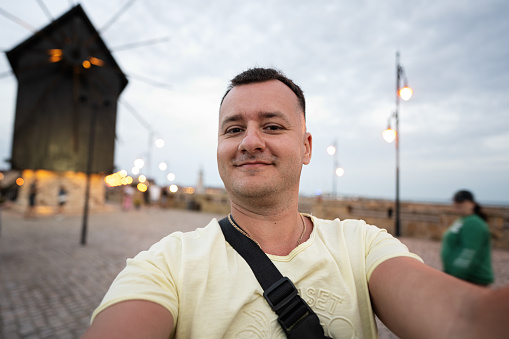 Portrait of a smiling tourist man taking selfie on the promenade near mill of Nessebar, Bulgaria.