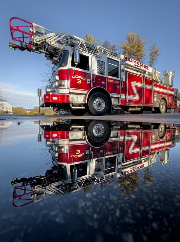 Brookline, United States – April 28, 2023: A close-up of a red fire truck is seen in the reflection of a large puddle of water with a bridge in the background