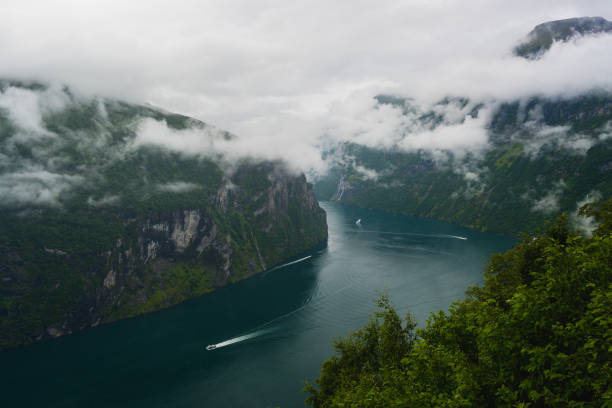 A horizontal shot from the Eagle Bend Lookout captures Geirangerfjord in Norway on a misty and rainy summer morning. Low clouds envelope the mountain peaks, and in the fjord's serpentine blue waters, three vessels are spotted. The mountainsides, blanketed with lush vegetation and rugged cliffs, frame this serene Nordic scene.