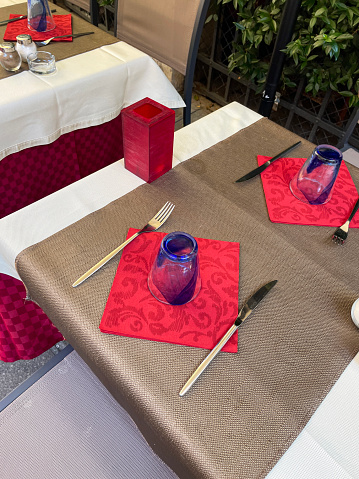 Stock photo showing close-up view of restaurant place settings at row of tablecloth covered tables outside a pavement cafe square.