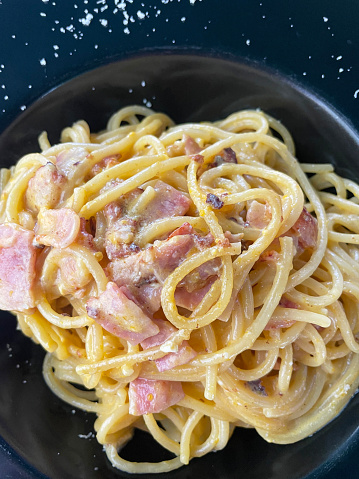 Stock photo showing close-up, elevated view of a black plate with a portion of spaghetti carbonara, using a traditional Italian recipe. The pasta is mixed with eggs, bacon lardons, grated Italian Parmesan cheese and garlic.