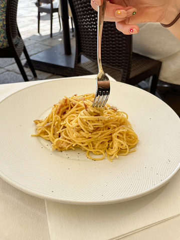 Stock photo showing close-up view of a white plate with a portion of spaghetti carbonara, using a traditional Italian recipe. The pasta is mixed with eggs, bacon lardons, grated Italian Parmesan cheese and garlic.