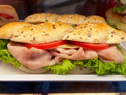 Stock photo showing close-up view of bakery display shelf containing row of ham and cheese salad submarine roll sandwiches, available for purchasing.