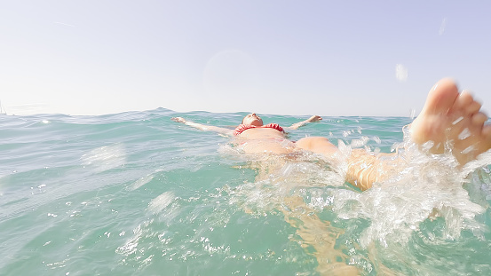 unrecognizable mature adult woman swimming on her back in the ocean water and splashing with her feet in summer. relax, wellbeing and wellness concept.