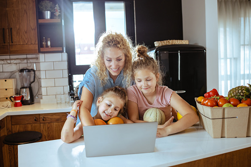 A mid adult Caucasian woman and her two young daughters are embracing while searching together for a healthy recipe on their laptop.