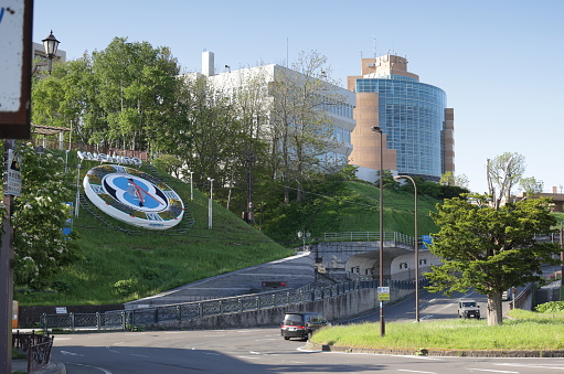 Kushiro City, Japan - June 6, 2023: A large clock face with a Kushiro sign rests on a grassy slope in Nusamai Park in the Nusamaicho district. The curved Kushiro City Museum of Art stands in the background. Traffic flows on Route 113 near the south side of the Kushiro River. Spring morning with clear skies over the port city by the Pacific Ocean.