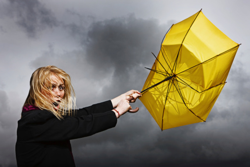 A pretty blonde woman looks horrified and helpless as her frayed and broken umbrella blows away from her, dragging her along, in a thunderstorm. 