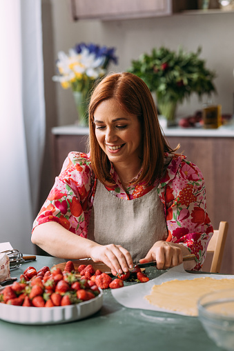 A smiling Caucasian female cutting strawberries while making a cake. She is sitting at the kitchen table.