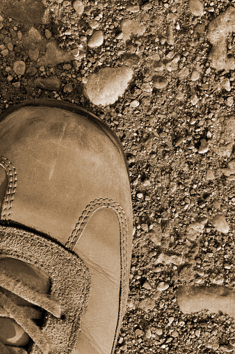 Hiking boot off-road olive green shoe on hard arid dried rough terrain ground soil, vertical flat lay, large detailed sepia macro closeup, barren earth, dust, stones, rocks, pebbles, sand background