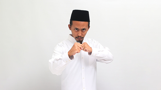 an Angry young Indonesian man wearing a songkok or peci or kopiah, is ready to fight, with a defensive fist gesture, feeling mad and upset, isolated on a white background