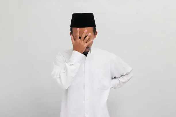 an Ashamed and embarrassed Young Indonesian man, wearing a songkok, peci, or kopiah, facepalms, covering his face with his hand, isolated on a white background