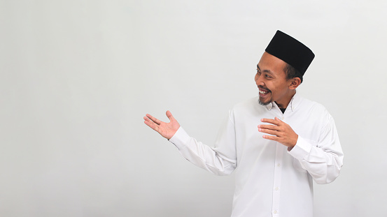 Excited Young Indonesian man, wearing a songkok, peci, or kopiah, extends his hands to the side of the copyspace, inviting someone to come, isolated on white background
