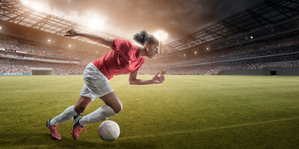 professional female soccer player running with ball in floodlit football stadium full of spectators at sunset.