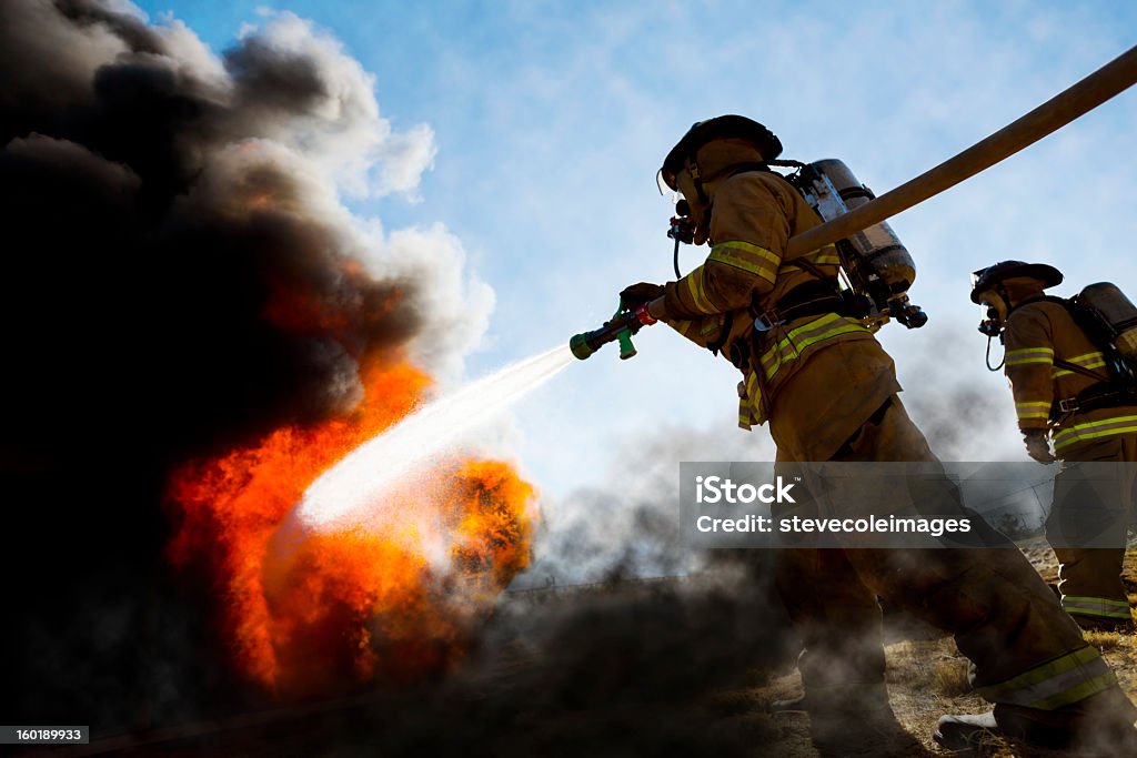 Firefighters Extinguishing House Fire Firefighters in a fire protection suit wearing firefighter helmet with breathing device and holding fire hose is extinguishing a burning house fire that is putting off excessive heat and smoke.  Fire could have been caused by accident or arson.  Second firefighter in background.   Firefighter Stock Photo