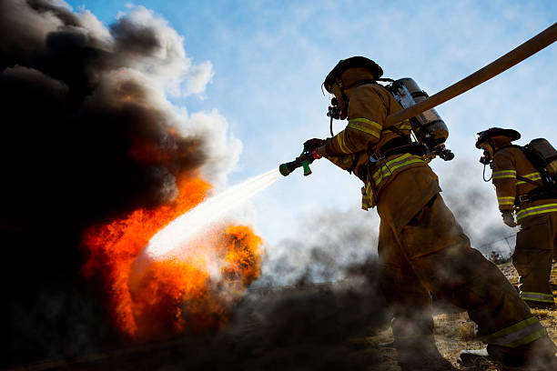 Firefighters Extinguishing House Fire Firefighters in a fire protection suit wearing firefighter helmet with breathing device and holding fire hose is extinguishing a burning house fire that is putting off excessive heat and smoke.  Fire could have been caused by accident or arson.  Second firefighter in background.   fire natural phenomenon photos stock pictures, royalty-free photos & images