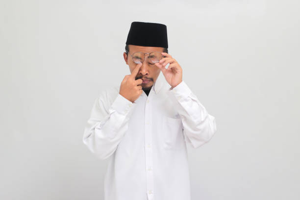 Young Indonesian man is experiencing sore eyes due to dust allergy and overwork A young Indonesian man, wearing a songkok, peci, or kopiah, is experiencing sore eyes and rubbing his swollen eyes due to dust allergy, itching and overwork, isolated on white background human eye scratching allergy rubbing stock pictures, royalty-free photos & images