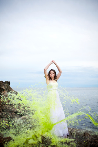 Dramatic moment of impact: green liquid paint thrown at/hitting a beautiful bride standing at the oceanfront with arms raised.