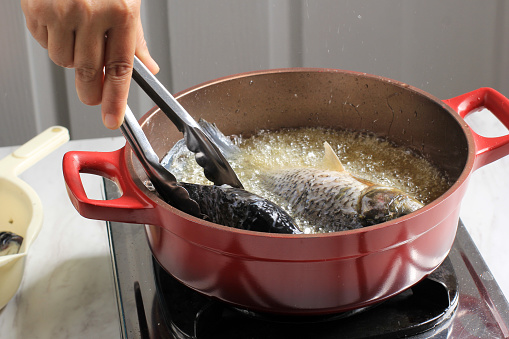 Female Hand using Stainless Tongs, Process of Frying River Carp Fish in a Red Frying Pan. Goreng Ikan Mas