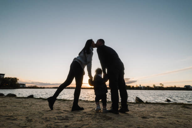 mother kisses father and daughter near lake in nature. silhouette at sunset. spending time together on beach on vacation. mom, dad hugs child and walk. concept of autumn holiday. family photo outdoors - silhouette kissing park sunset imagens e fotografias de stock