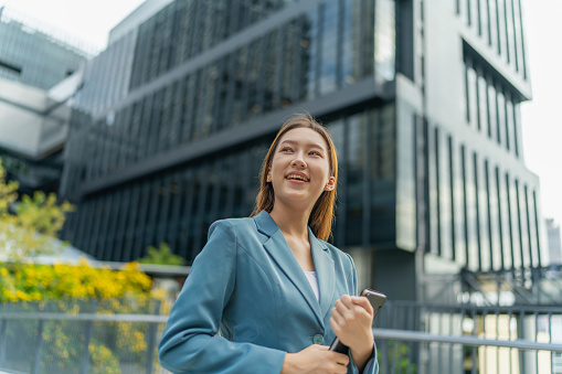 Exuding women power and leadership, the young Asian businesswoman stands before a modern contemporary office building, holding a digital tablet with a confident smile, embodying a tech-savvy symbol of her professional prowess