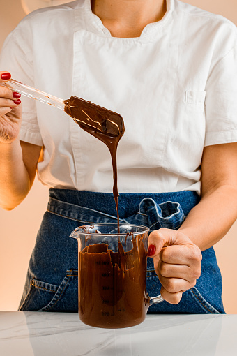 Woman in cooking uniform mixes melted chocolate and peanuts in transparet measuring cup with spatula. Ingredients for decorating desserts or cakes