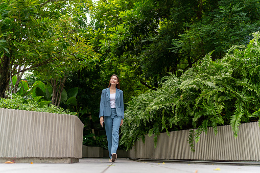 With a phone in hand, the young Asian businesswoman walks along a garden path, radiating happiness and vitality, seamlessly blending work and contentment amidst nature's beauty.
