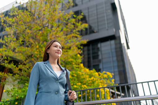 Radiating success and woman power, the young Asian businesswoman stands before contemporary corporate buildings, embodying empowered female leadership in the bustling cityscape of business on the go.