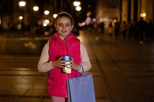 Against the backdrop of a bustling cityscape at night, a young daughter immerses herself in a moment of pure bliss, clutching a bag of warm popcorn and relishing the flavors as she explores the enchanting city street