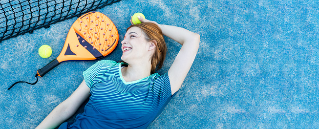 A pretty female paddle tennis player is lying on the grass while looking happily at a paddle tennis racket. Outdoor paddle tennis concept, women playing paddle tennis.