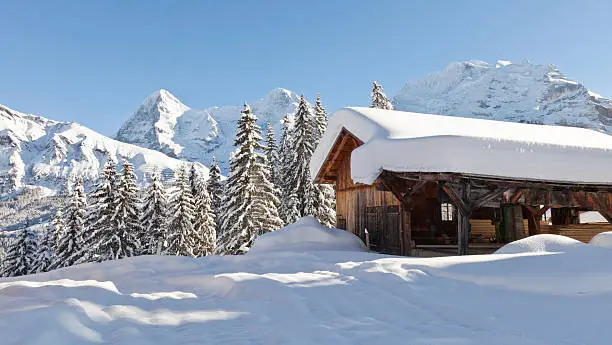 A wooden building is covered with heavy snow, with the Eiger, Mönch and Jungfrau mountains behind,