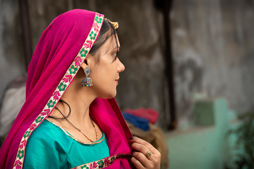 Portrait of a beautiful, traditional rural Indian woman in north Indian attire and covering her head with dupatta and looking away while thinking.
