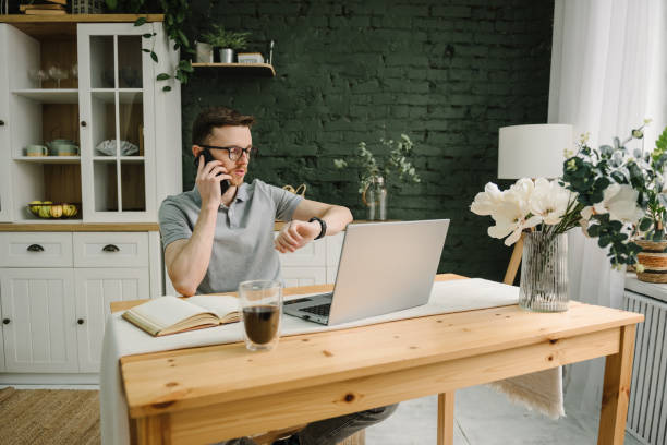 Coffee break time. Serious man working, learning online on laptop in home office. Trading cryptocurrency. Trader talk on phone and looks at his wristwatch, investor broker analyzing indexes. Freelance