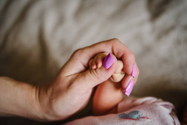 babyhand. junge mutter, die hände des kindes hält. nettes kleines baby, das zu hause auf dem bett liegt. mama hält sohn finger. closeup. winzige hand eines neugeborenen mädchens oder jungen, der in hände gewickelt ist. niedliche schlafende tochter. - furniture barefoot little boys family stock-fotos und bilder