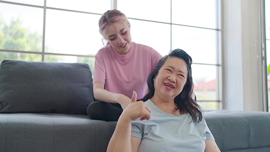 Asian daughter giving shoulder massage to elderly mother while relexing in living room at home. Young daughter giving an elderly mother relaxing massage. Family lifestyle