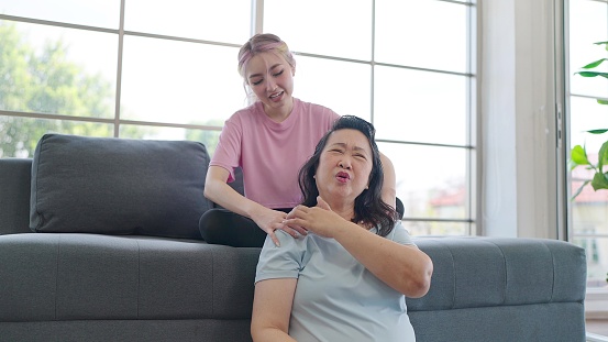 Asian daughter giving shoulder massage to elderly mother while relexing in living room at home. Young daughter giving an elderly mother relaxing massage. Family lifestyle