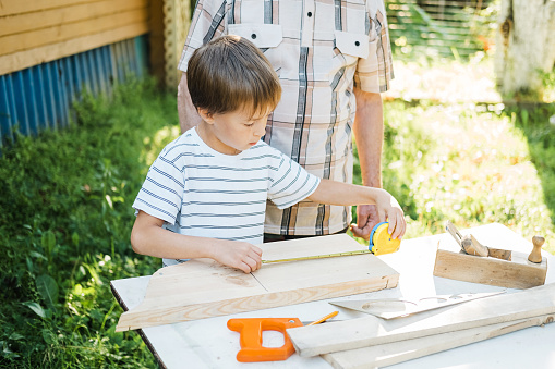 An elderly gray-haired older man and a small boy are standing at a table with carpentry tools. Grandfather teaches his grandson to carpenter in the garden on a sunny day