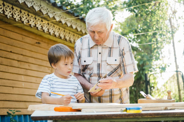 istock An elderly gray-haired older man and a small boy are standing at a table with carpentry tools. Grandfather teaches his grandson to carpenter in the garden on a sunny day. 1602833821