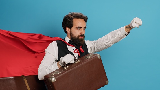 Hotel bellhop acting as superhuman with cape, offering to help people with suitcase luggage in studio. Young adult doorman presenting comic hero character with red fabric, feeling determined.