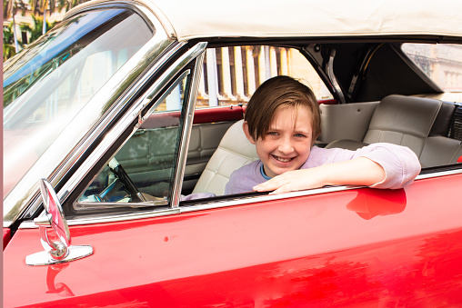 happy boy sits in the driver's seat of a red retro car. joyful 10 year old child driving a car, looks out of the window.