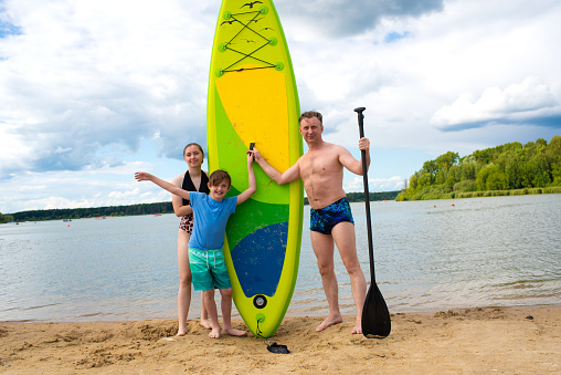 family posing with supboard on the background of the lake. a teenage girl, a little boy and her father spend the weekend in nature, plan to ride a stand-up board.