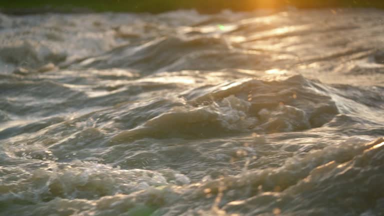 Afternoon sun shines on rapid river waves, drops of water glowing in air, closeup slow motion video