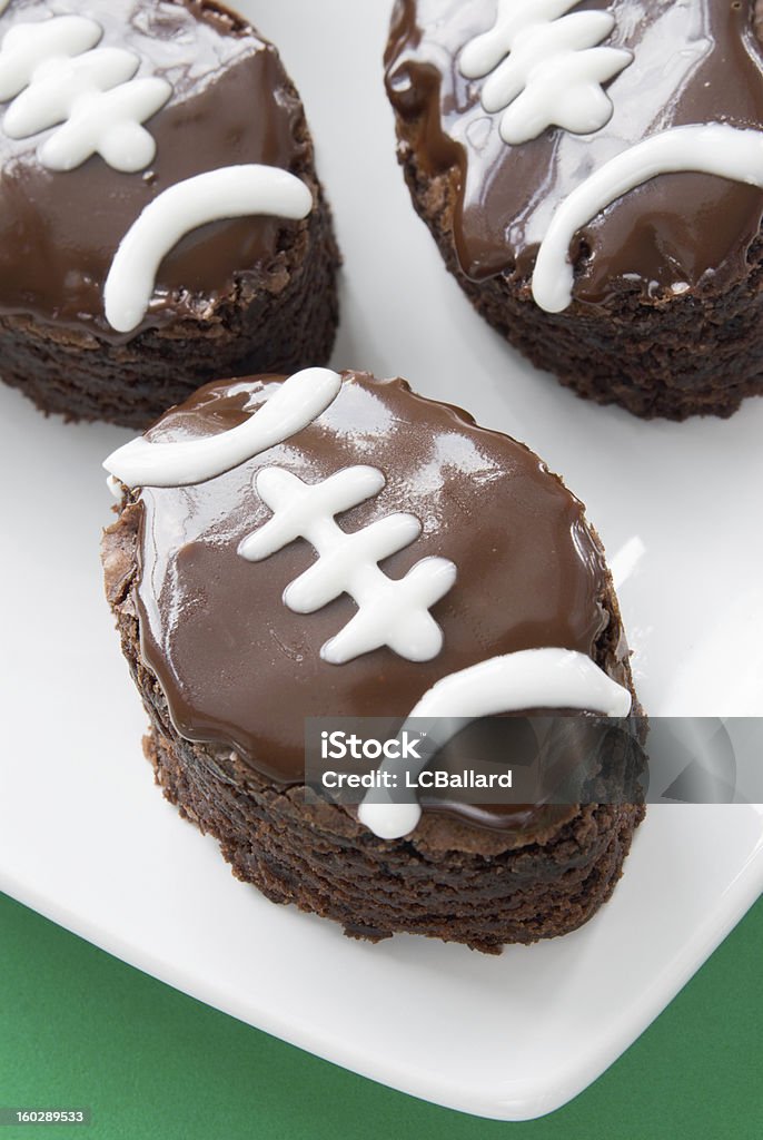 Three football shaped double fudge chocolate brownies on white plate Three homemade football shaped double fudge chocolate iced brownies with white piping that makes the brownies look like a football. Shallow definition of field. The brownies are served on a square white plate with a bright green paper background. American Football - Sport Stock Photo