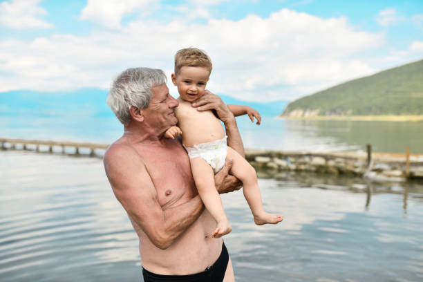 해변에서 휴가를 보내는 동안 호수 근처에서 귀여운 아기를 안고 있는 할아버지 - grandson water waterfront portrait 뉴스 사진 이미지