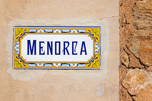 A hand painted Spanish tiled sign panel with painted Menorca text with a decorative Spanish motif border, inset into a distressed paint wall on the island of Menorca in the Balearic Islands, in the Mediterranean. Good copy space.