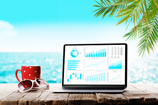Laptop business reports and charts and coffee cup on wooden table in front of sunny sea and palm leaves. Work and travel or remote business concept